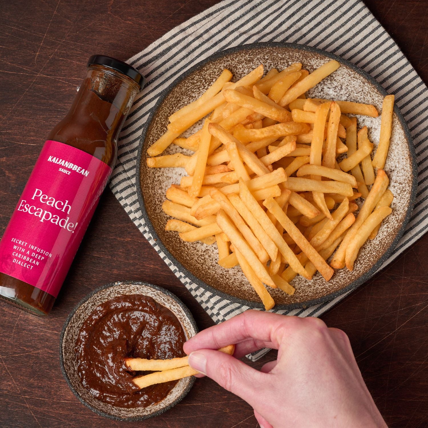 Plate of fries with a bottle of 'Peach Escapade' sauce on a wooden table.