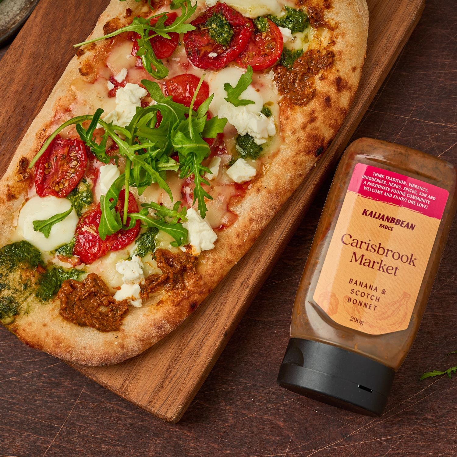 Pizza with tomatoes and arugula on a wooden board next to a bottle of Kalamazoo Carisbrook Market jam.